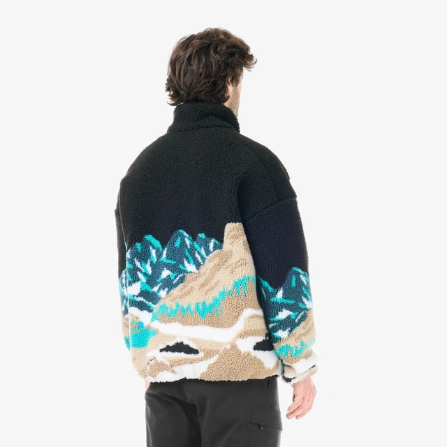 A mans stands in a white studio space wearing black pants and a black fleece with a mountain scape design on the bottom half of the fleece