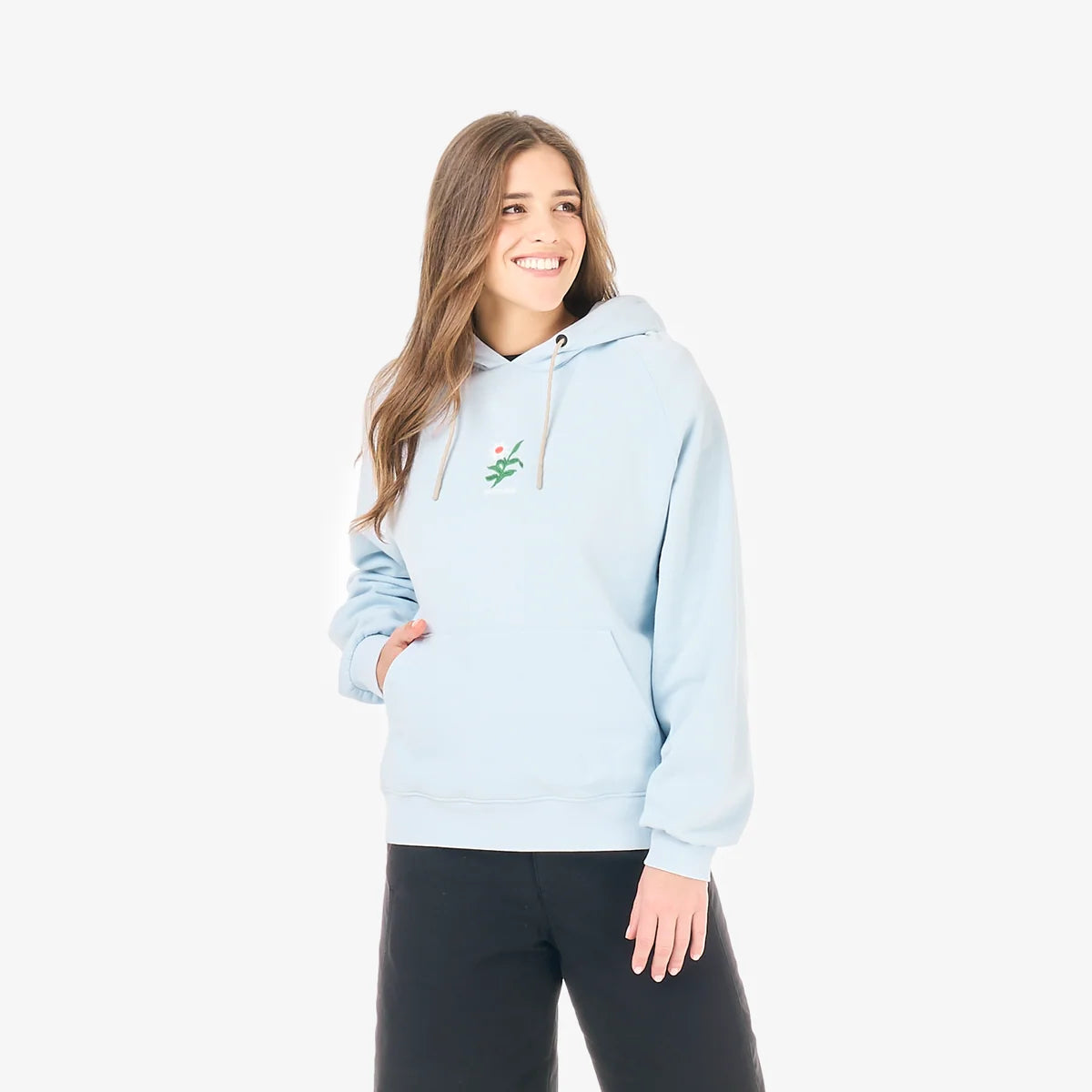 A woman stands in a white studio wearing a blue hoodie with a flower design on the front and black pants