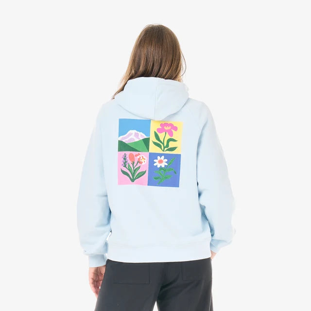 The back view of a woman stands in a white studio wearing a blue hoodie with a flower design on the front and black pants