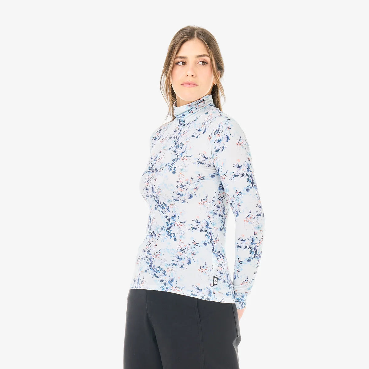 A woman stands in a white room wearing a floral print mock neck long sleeve and dark pants 