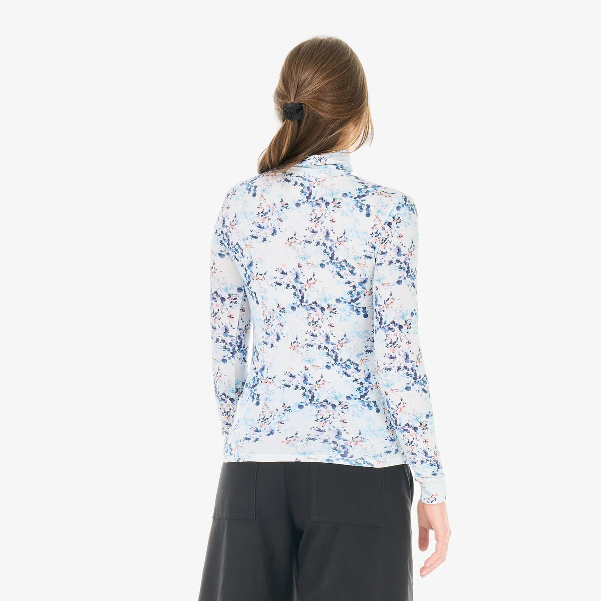 The back view of a woman standing in a white studio space wearing a floral print mock neck long sleeve and dark pants  