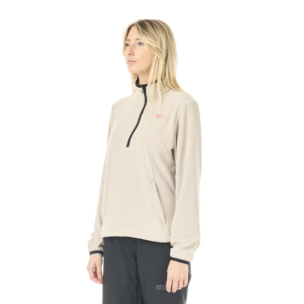A woman stands in a white studio space wearing a cashmere coloured 1/4 zip top and black pants