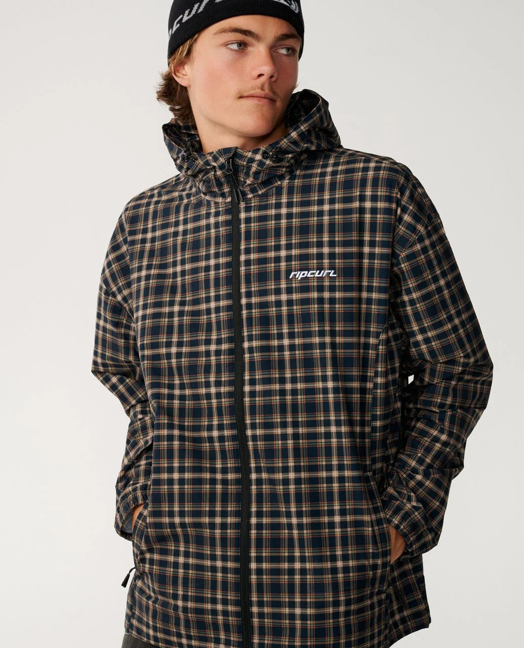 Rip Curl Archive DWR Plaid Jacket A man wearing a dark brown and black plaid hooded jacket with a white 'ripcur' logo on the chest, paired with a black beanie, against a plain light gray background.