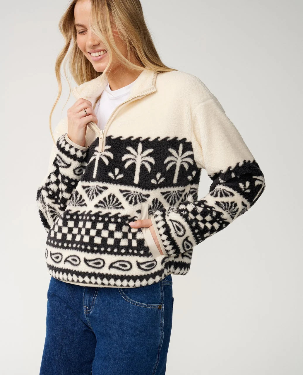 A woman stands in a studio wearing a black and white 1/4 zip fleece and jean