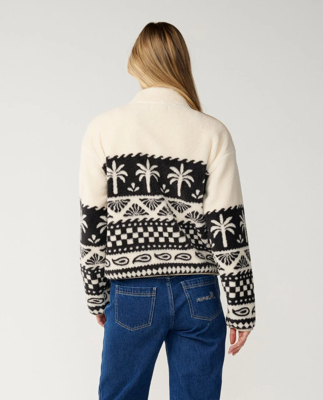 The back view of a woman in a white studio wearing a black and white fleece sweater and blue jeans