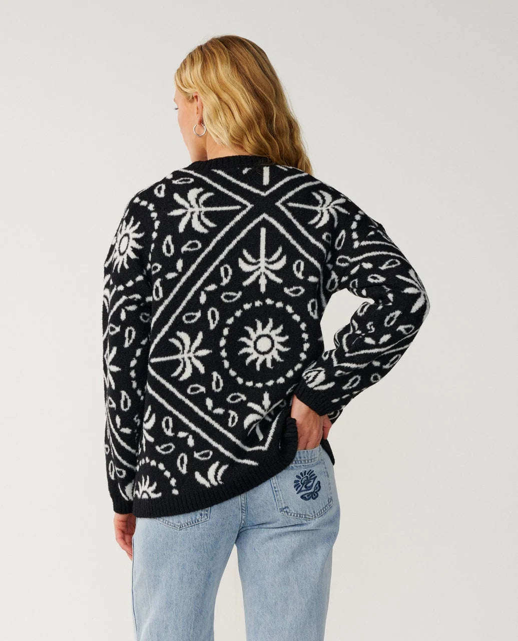 A woman stands in a white studio with her hand in back pocket wearing jeans and black and white knit sweater from Rip Curl