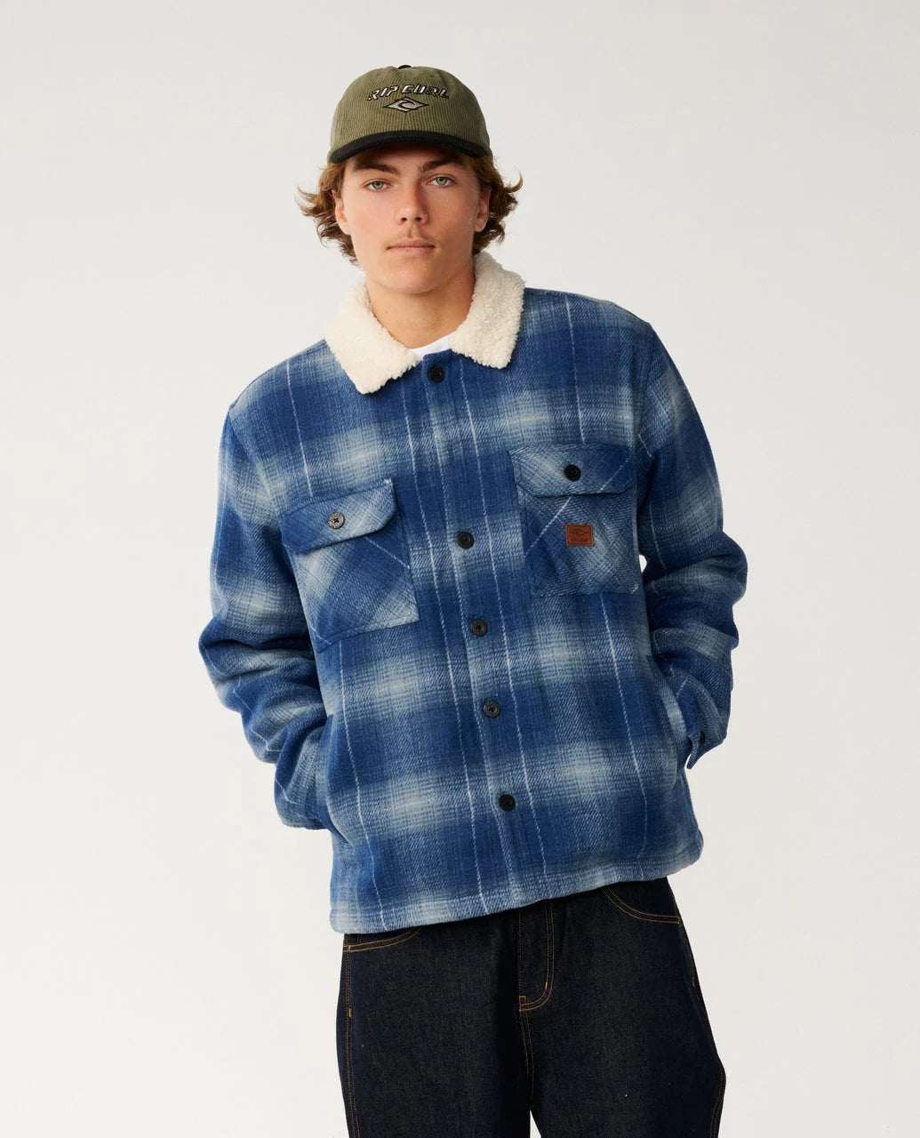 A man stands in a white studio wearing a blue plaid fleece lined jacket and dark pants