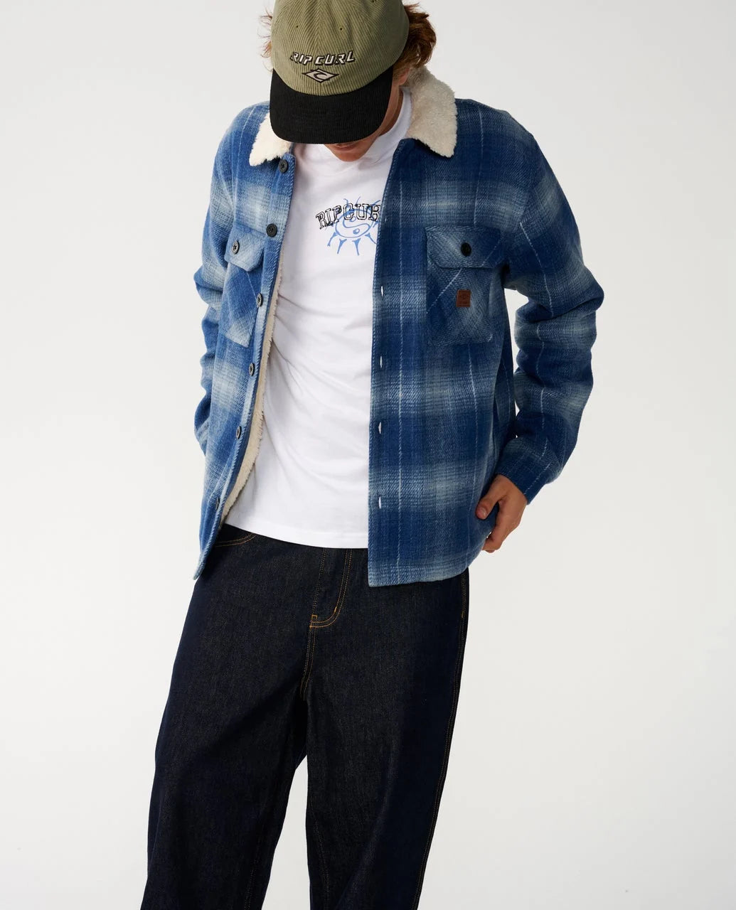 A man stands in a white studio wearing a blue plaid fleece lined jacket unbuttoned and dark pants