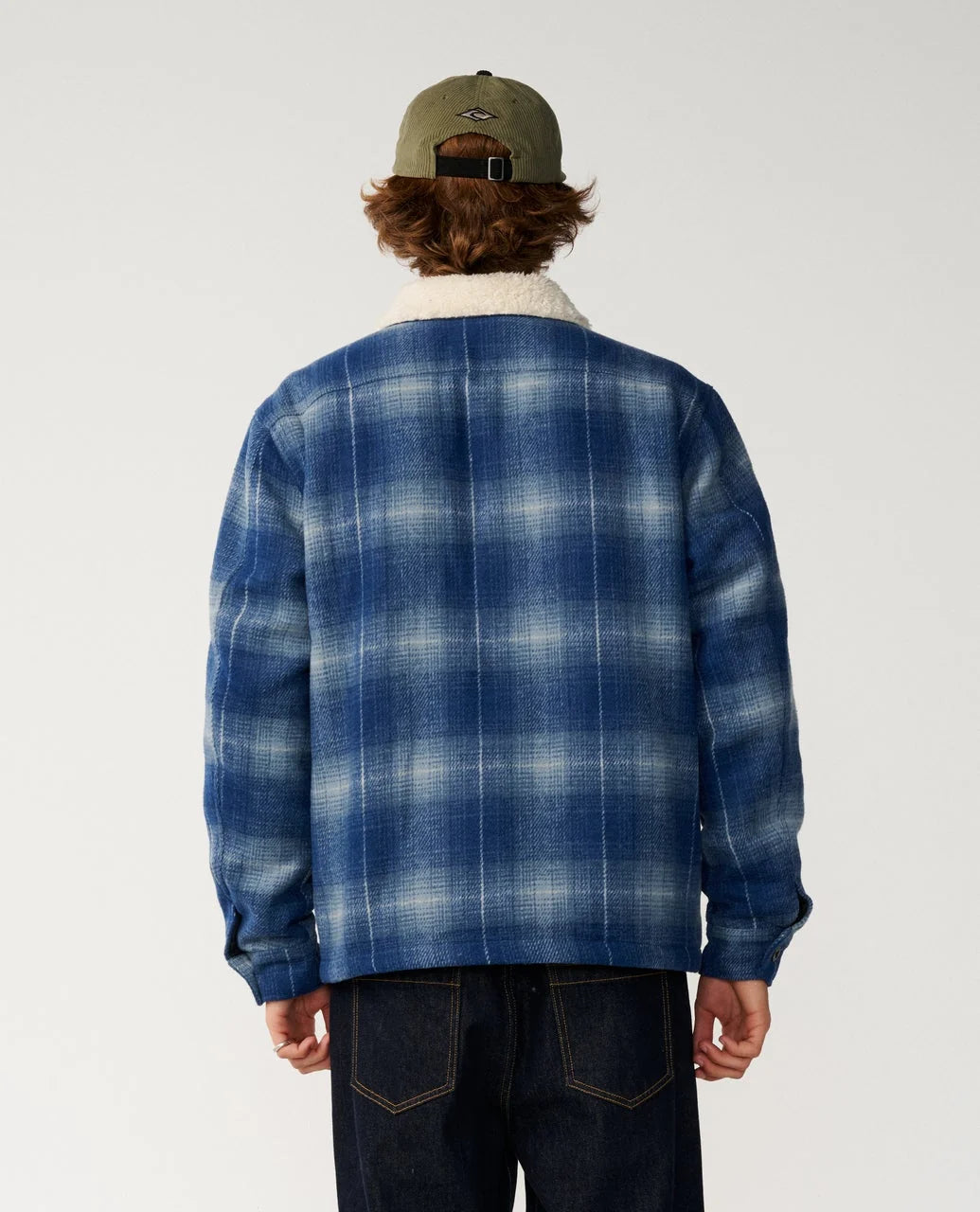 The back view of a man standing in a white studio space wearing a  blue plaid fleece lined jacket