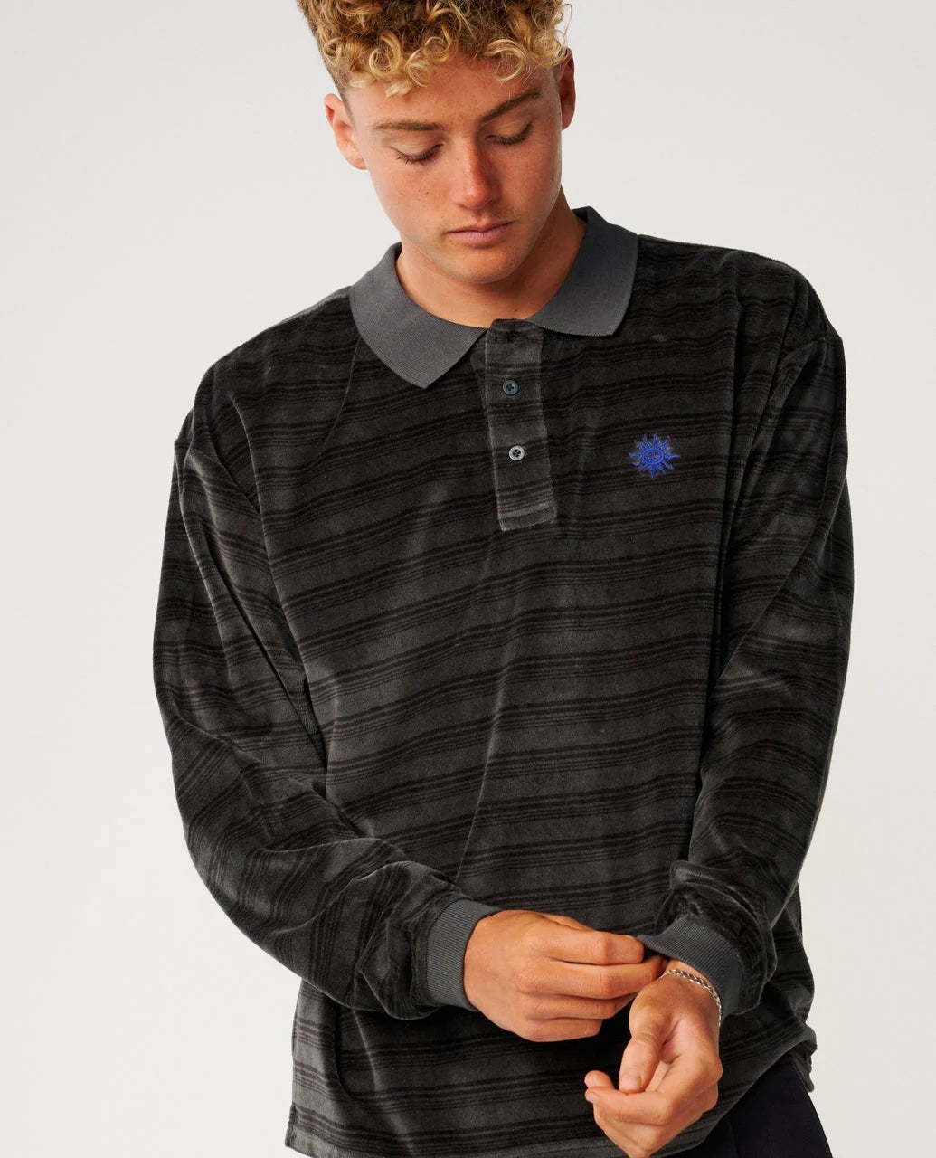 A man stands in a white room wearing a black striped polo from Rip Curl
