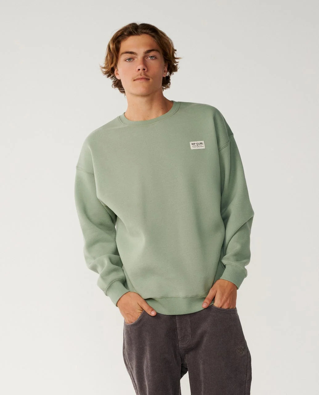  A man stands with his hands in his pockets wearing dark pants and green Rip Curl Crew Sweater