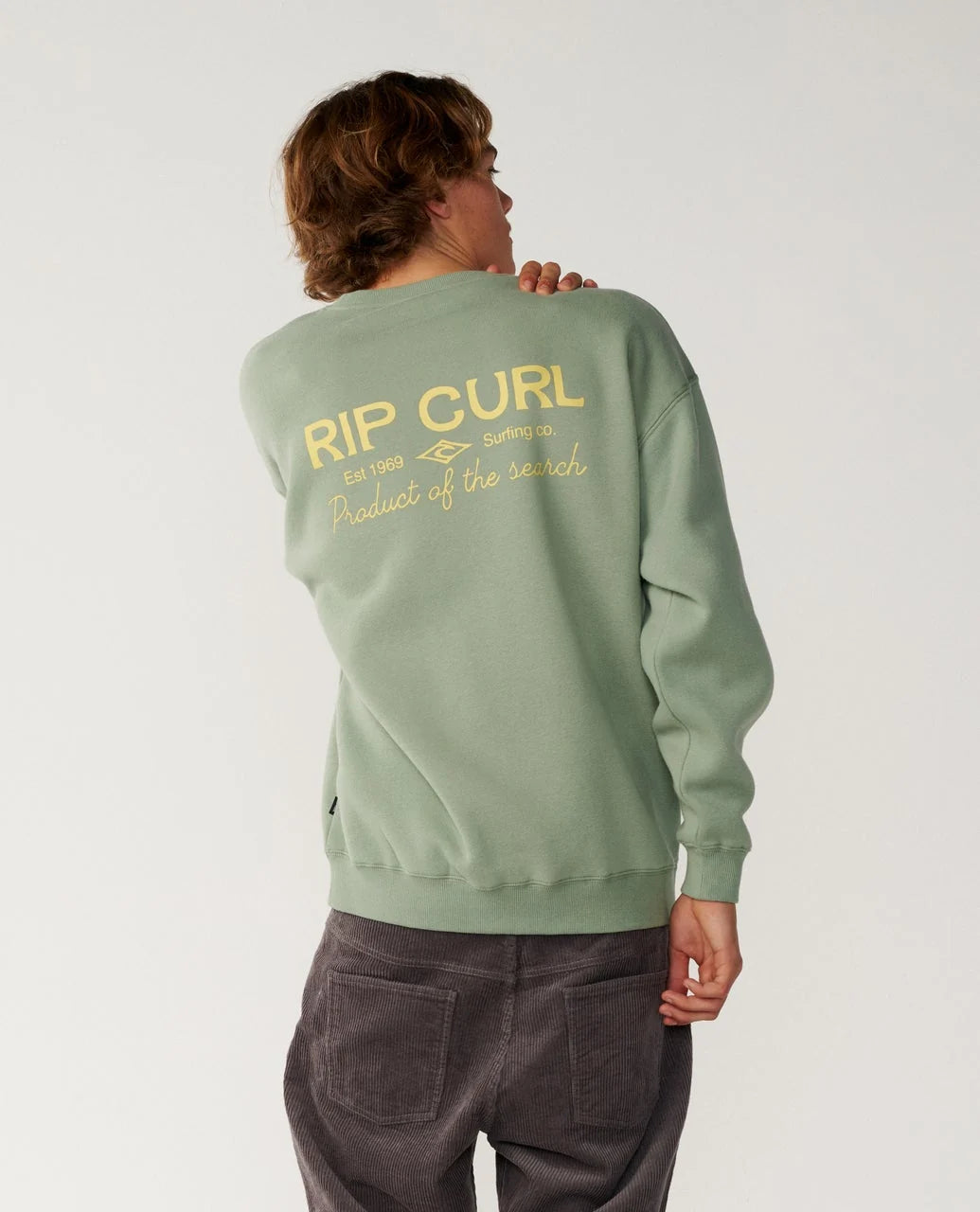 The back view of a man wearing a rip curl sage green crew sweater and dark pants.