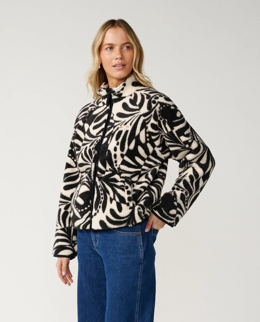 A woman stands in a studio space wearing a full zip Rip Curl fleece with black and white pattern and jeans