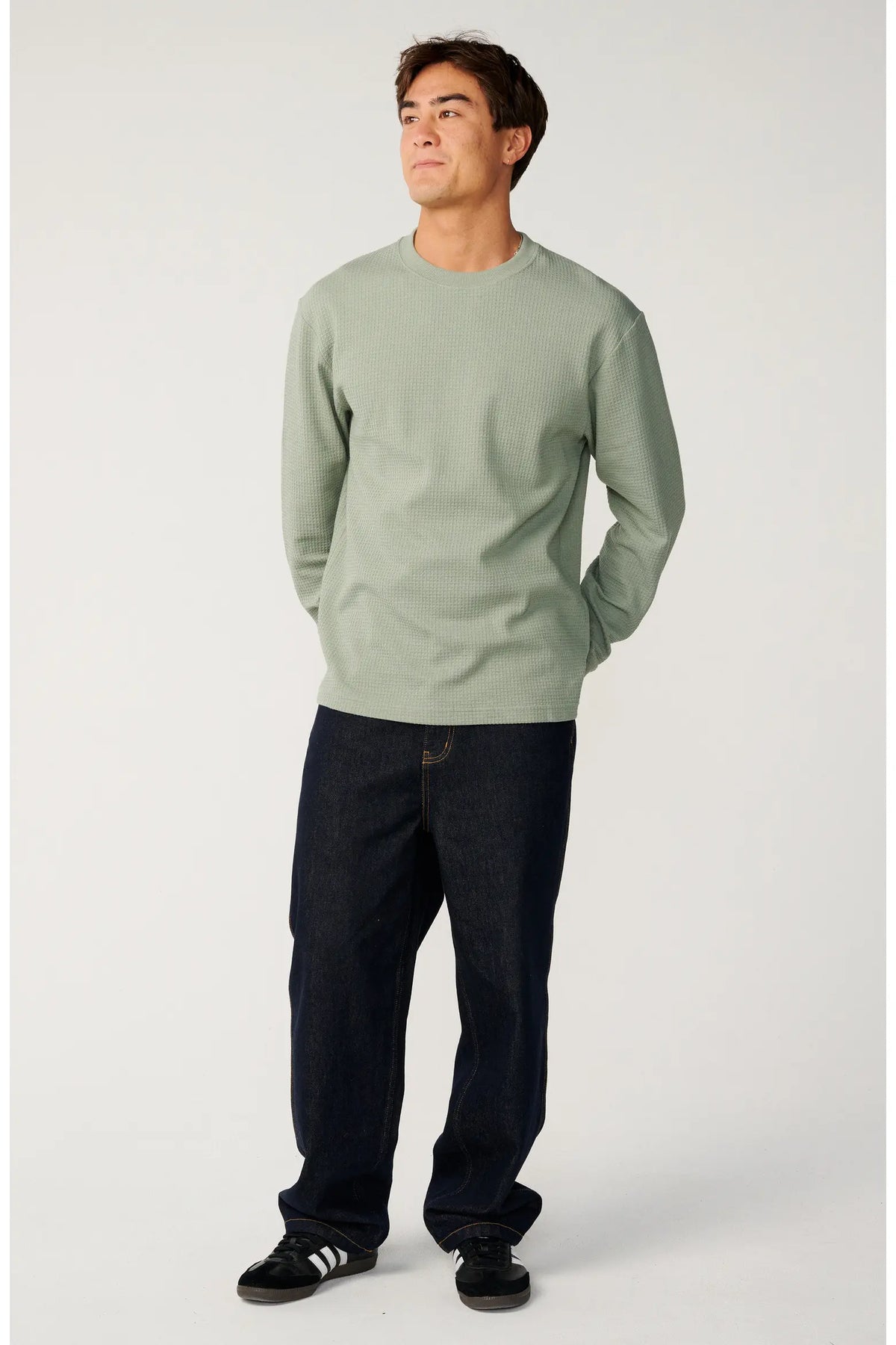 A man wearing a sage green long-sleeve crewneck sweatshirt, dark blue jeans, and black sneakers with white stripes, standing against a light gray studio background.