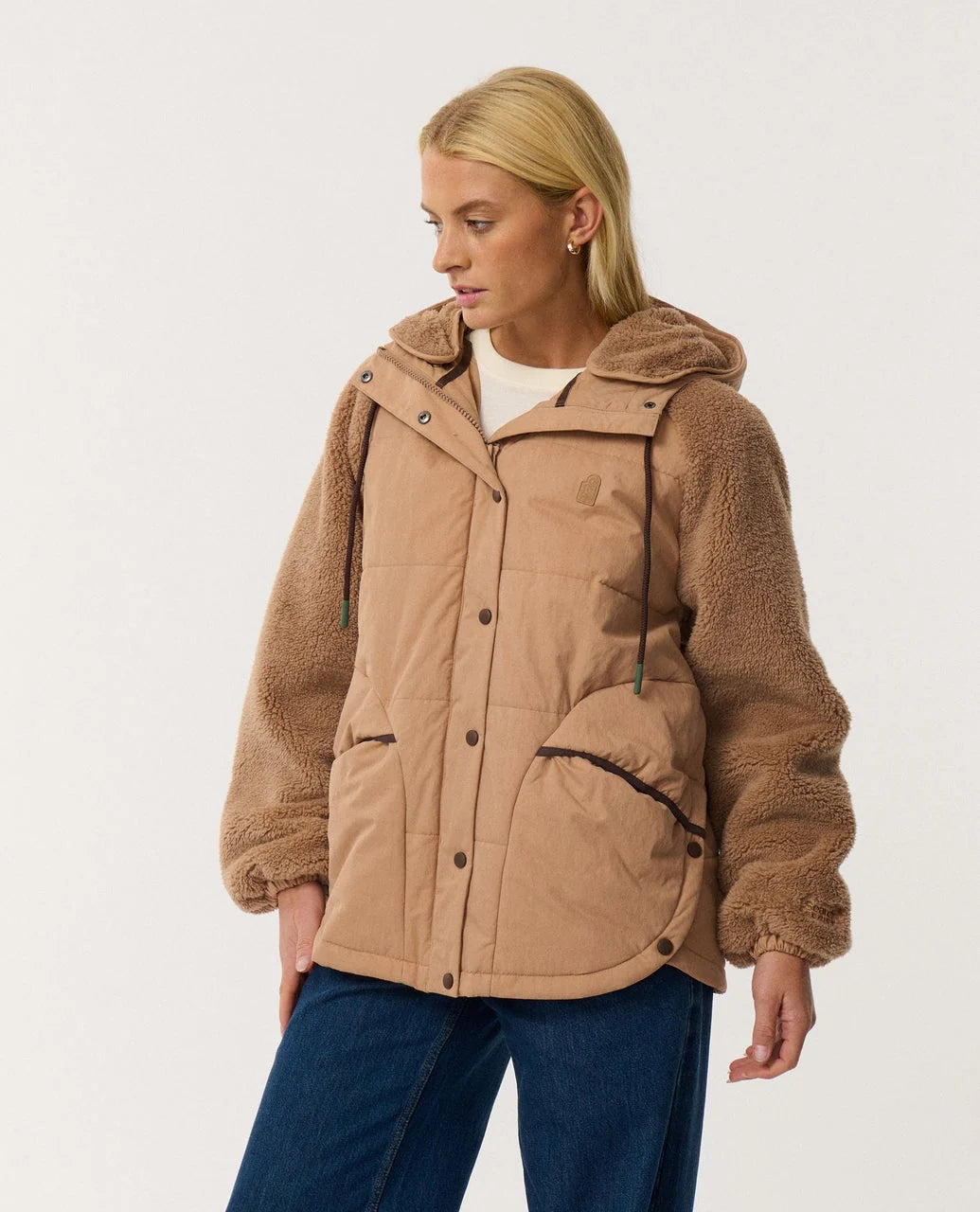 A woman stands in a studio space wearing a brown two tone button up jacket with fleece arms