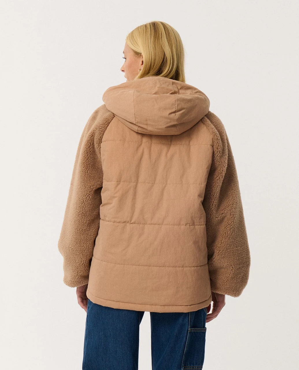 The back view of a woman standing in a white studio space wearing brown jacket