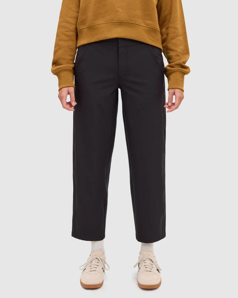 Tentree Breton Pant A person wearing black cropped trousers, a mustard yellow sweatshirt, cream socks, and beige sneakers, shown from the chest down against a light gray studio background.