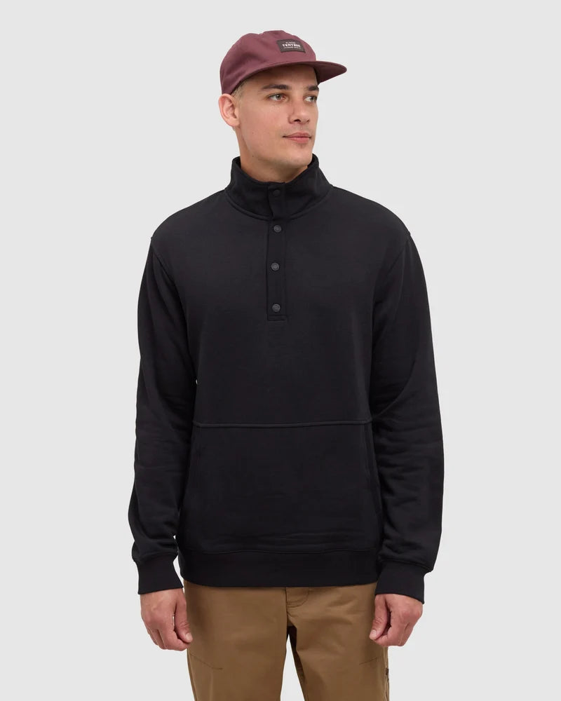 A man stands in a white studio space wearing tan pants and a black 1/4 snap top and a hat from Tentree