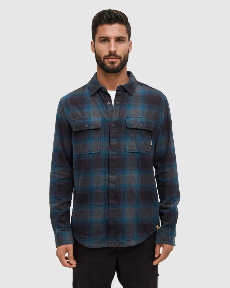 A man stands in white studio space wearing a blue plaid kapok shirt and dark pants