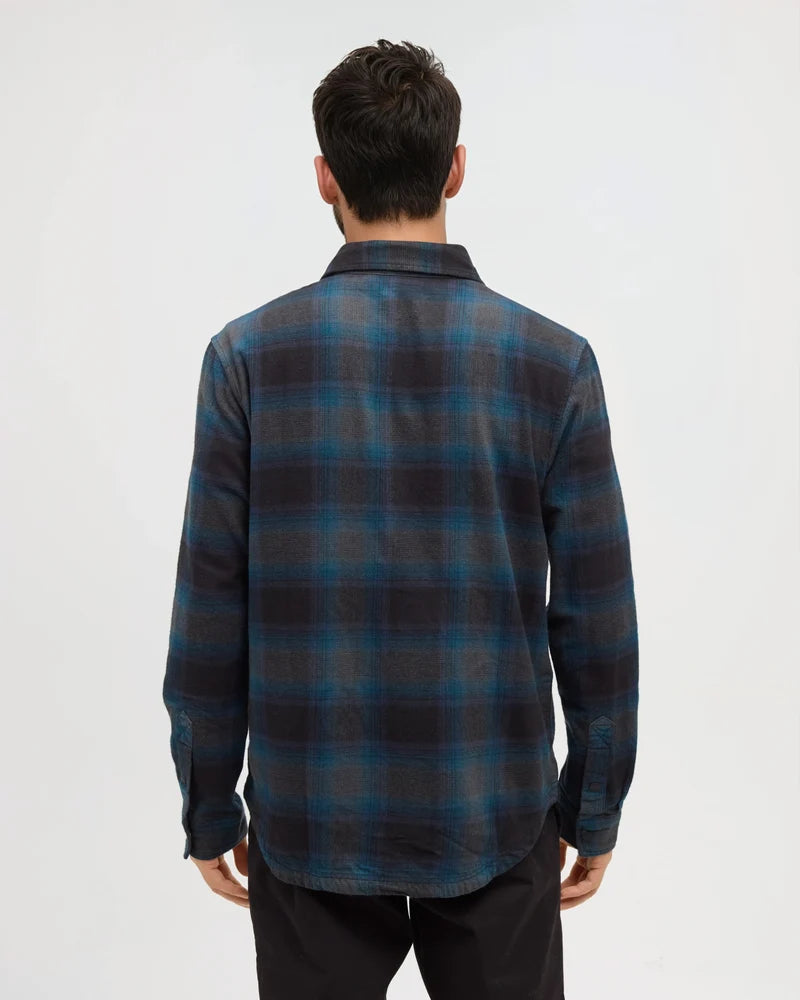 The back view of a man standing in a white studio space wearing a blue plaid shirt.