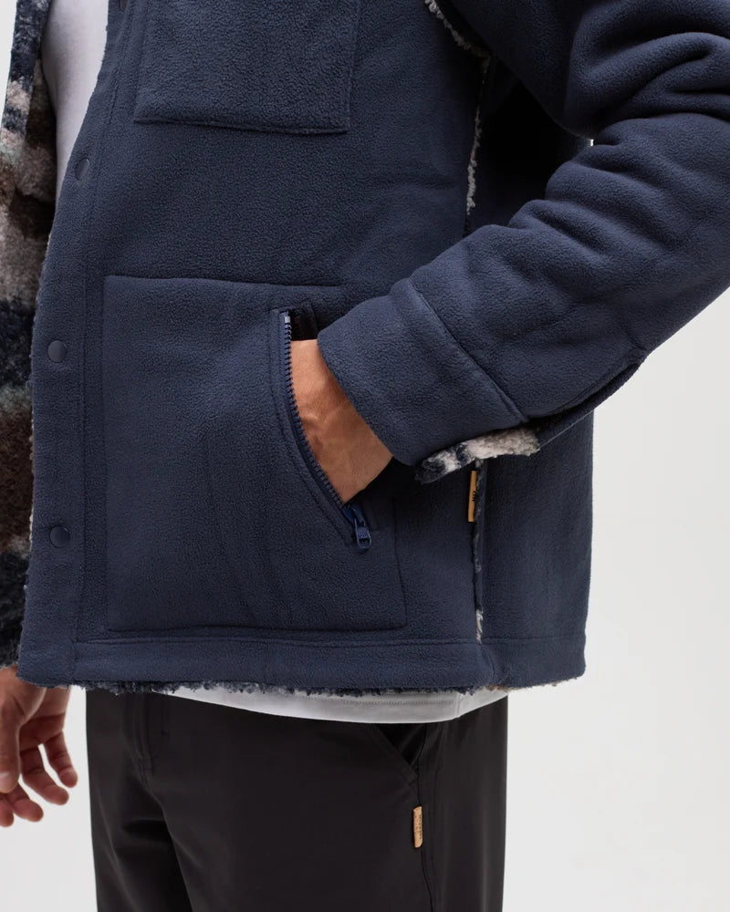 A man stands in a white studio space wearing a reversible blue jacket with his hand in his pocket.