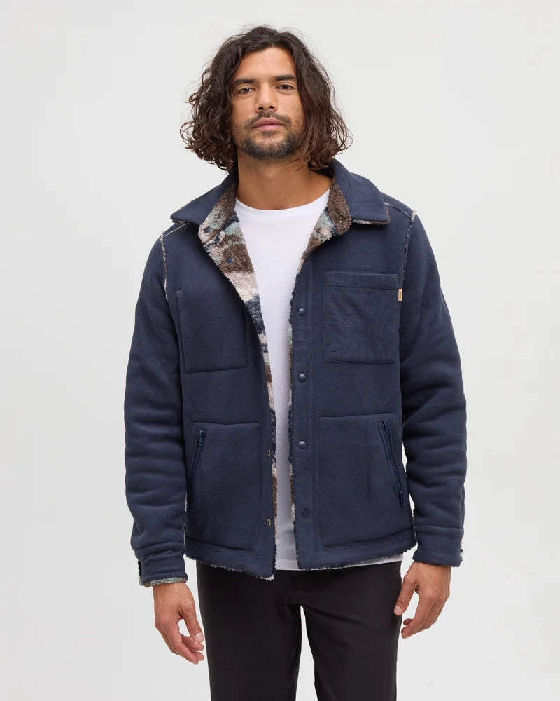A man stands in a white studio space wearing a reversible blue jacket and dark pants