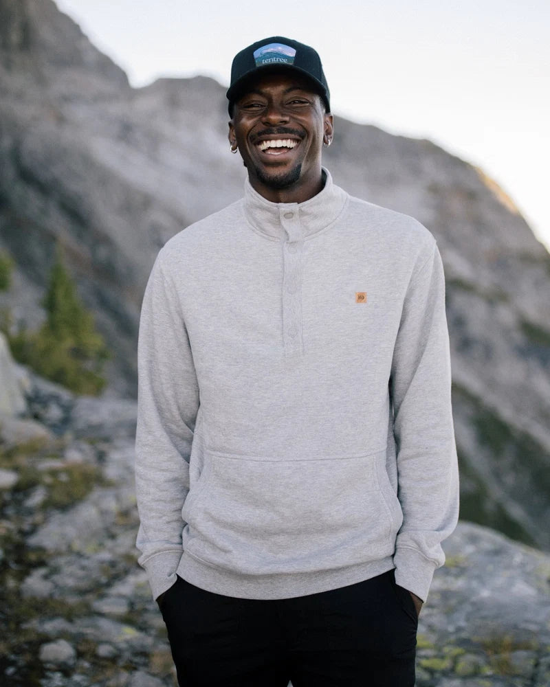Tentree Parks Souvenir Elevation Hat A smiling man wearing a light gray quarter-zip pullover sweatshirt, black pants, and a black baseball cap stands outdoors in front of rocky mountains.