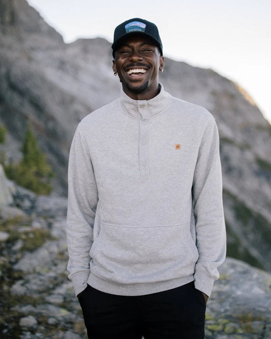 Tentree Parks Souvenir Elevation Hat A smiling man wearing a light gray quarter-zip pullover sweatshirt, black pants, and a black baseball cap stands outdoors in front of rocky mountains.