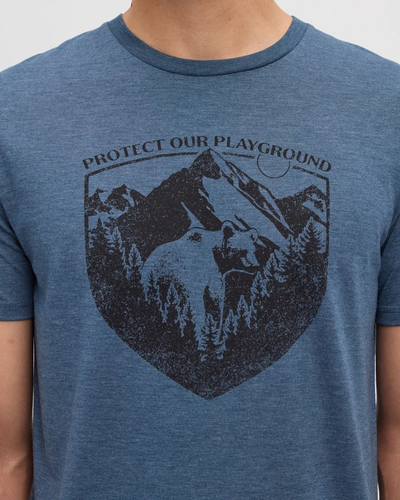 Tentree Protect Tee A close-up of a person wearing a heathered blue crewneck t-shirt with a dark graphic print on the front showing a mountain, bear, and pine trees, with the text 'PROTECT OUR PLAYGROUND'.