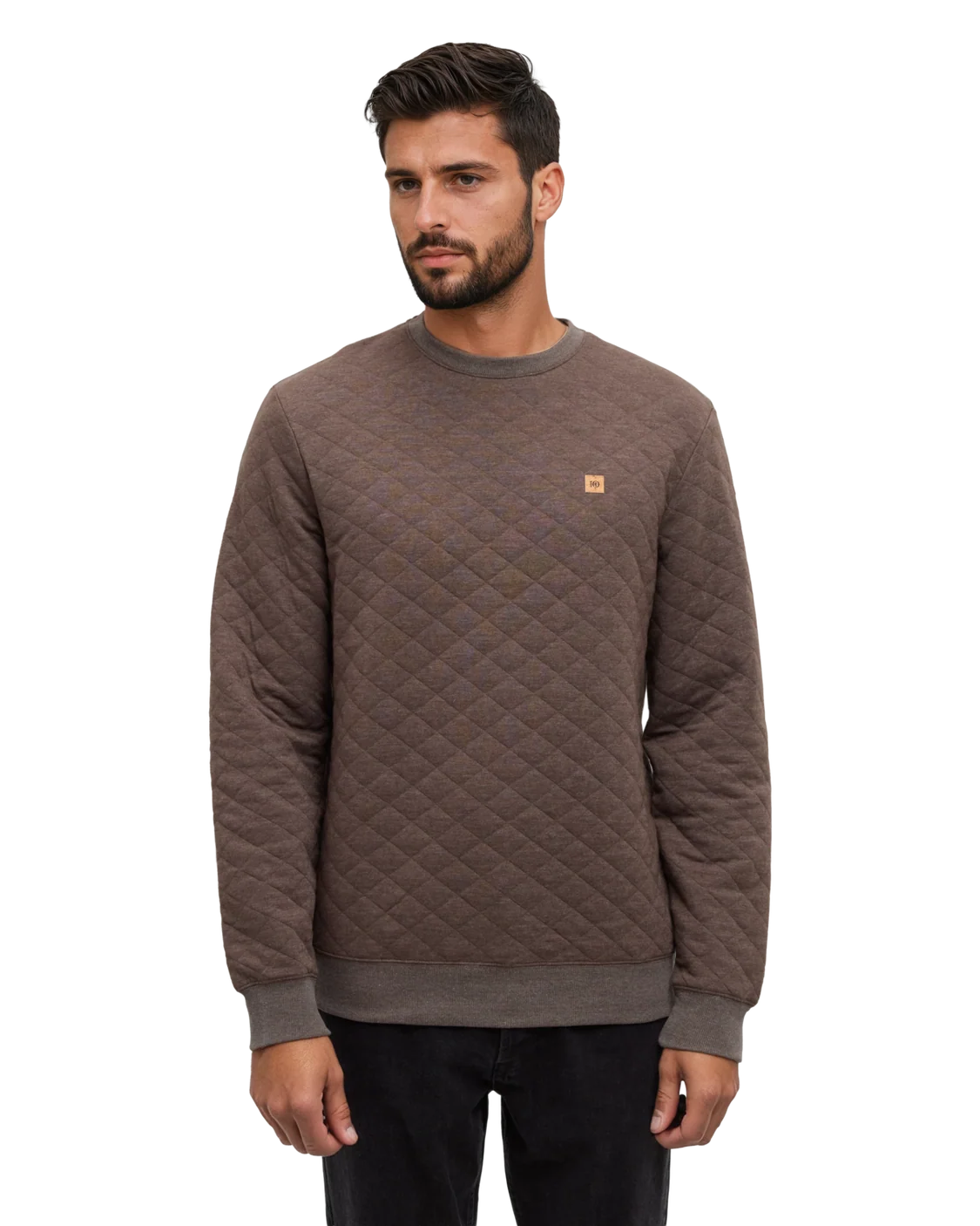 A man stands in a white studio space wearing a quilted brown sweater and dark pants.