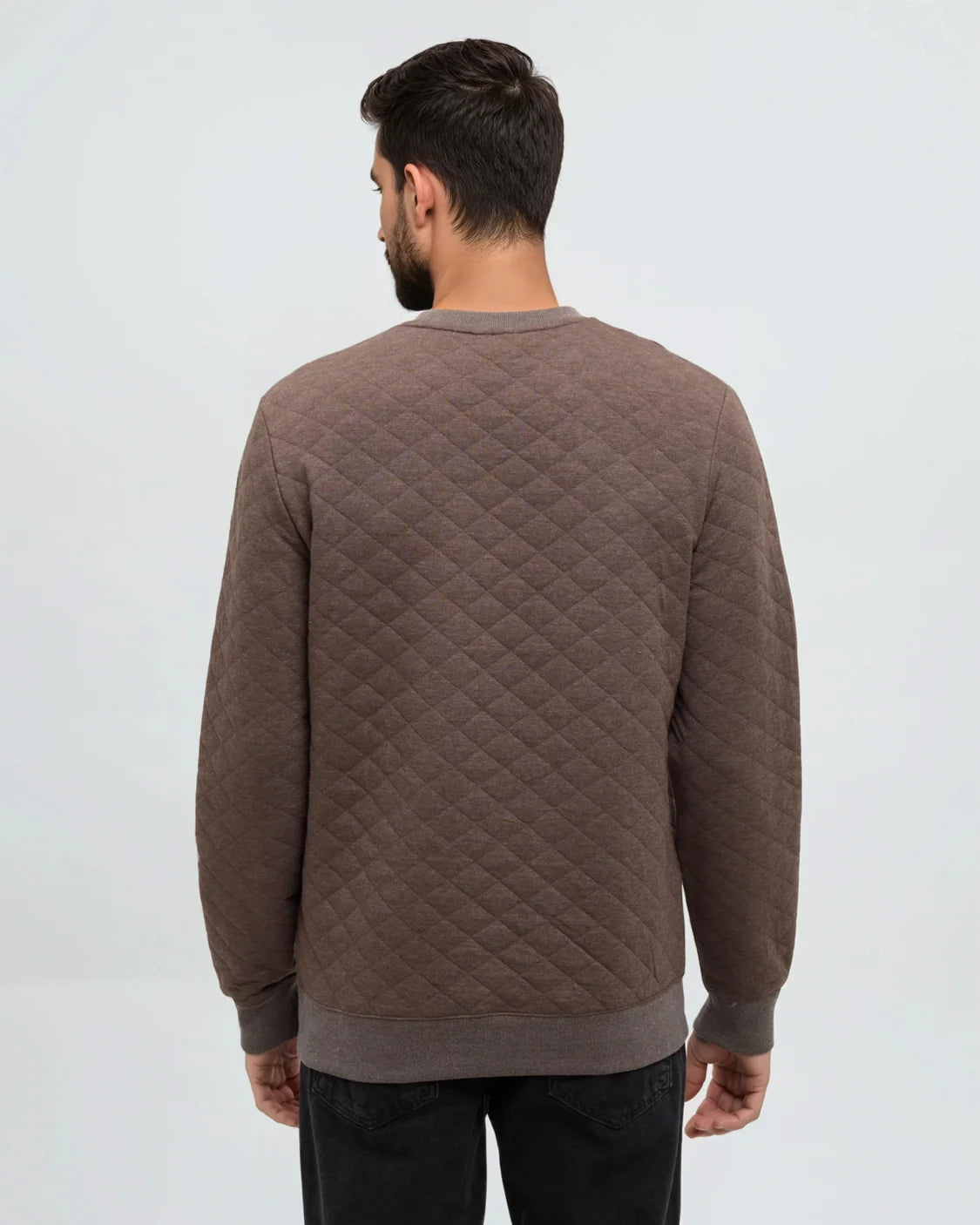 A view of the back of a man standing in a white room wearing a brown quilted Tentree sweater and dark pants