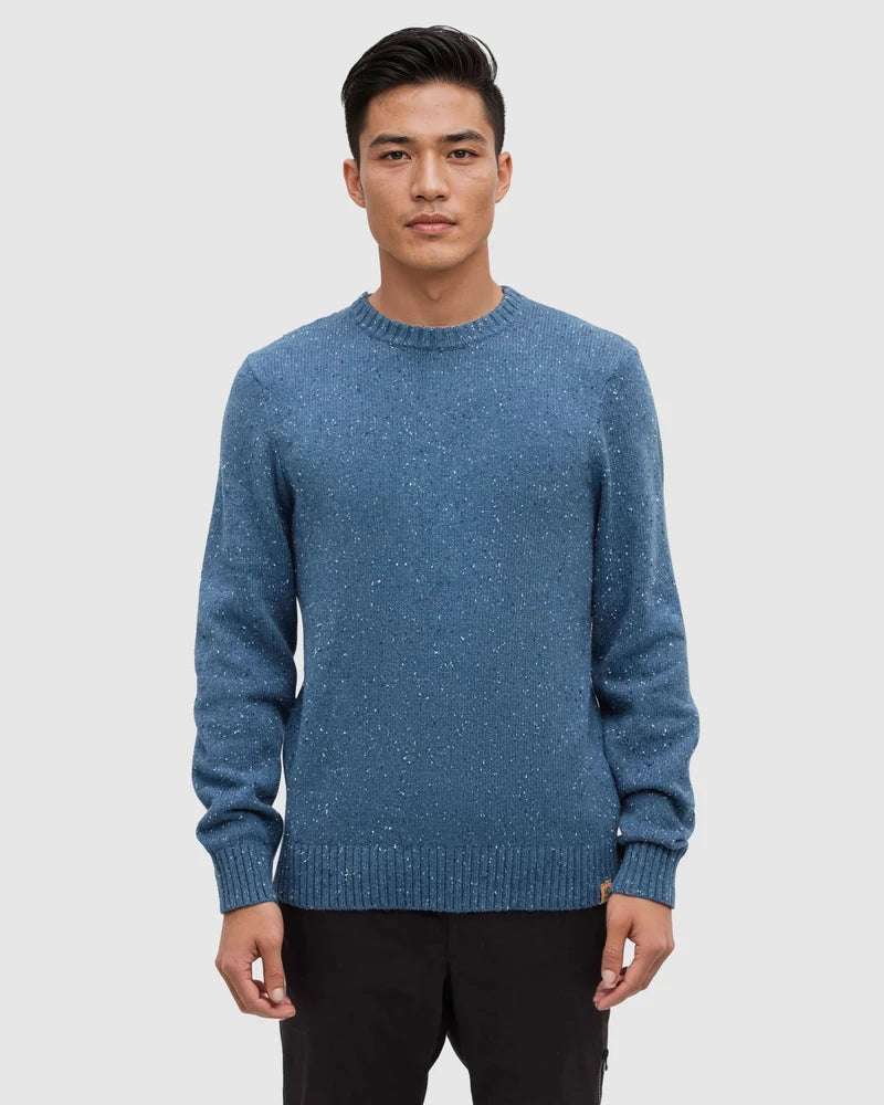 A man stands in a white studio space wearing a blue fleck rambler sweater and dark pants