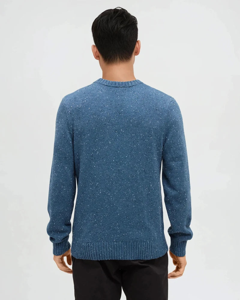 The back view of a man wearing a blue fleck sweater standing in a white studio space 