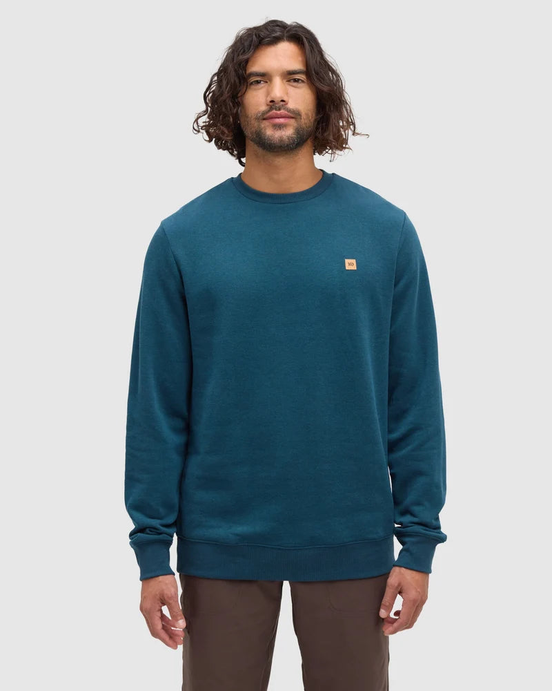 A man stands in a white studio space wearing a teal coloured crew sweatshirt from tentree