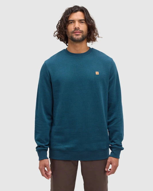 A man stands in a white studio space wearing a teal coloured crew sweatshirt from tentree
