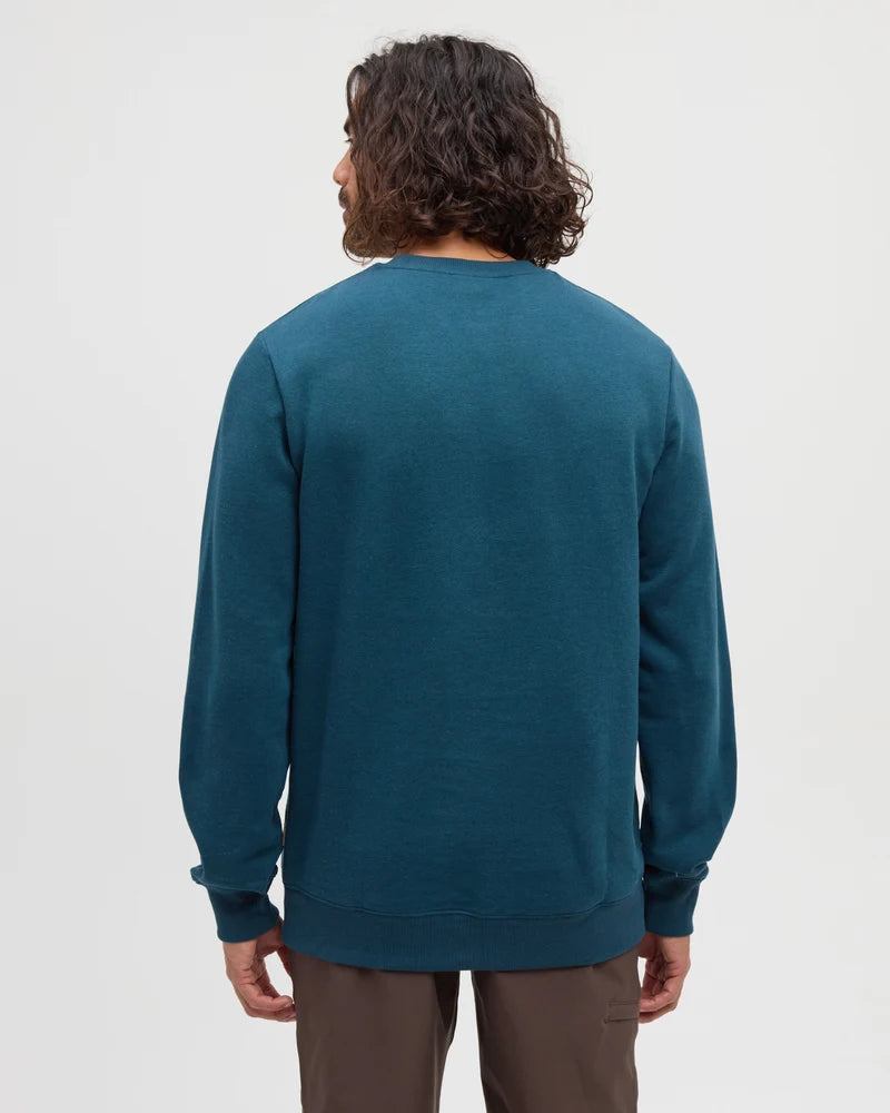 The back view of a man standing in a white studio space with a teal Reynard sweater and tan pants