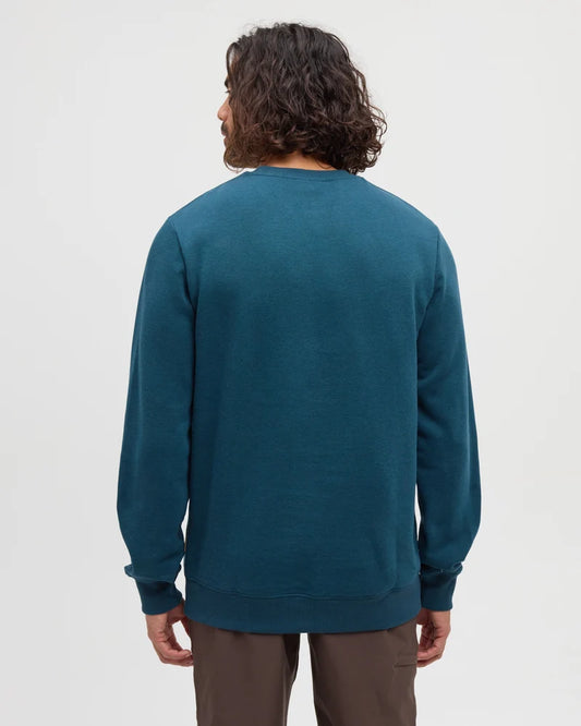 The back view of a man standing in a white studio space with a teal Reynard sweater and tan pants