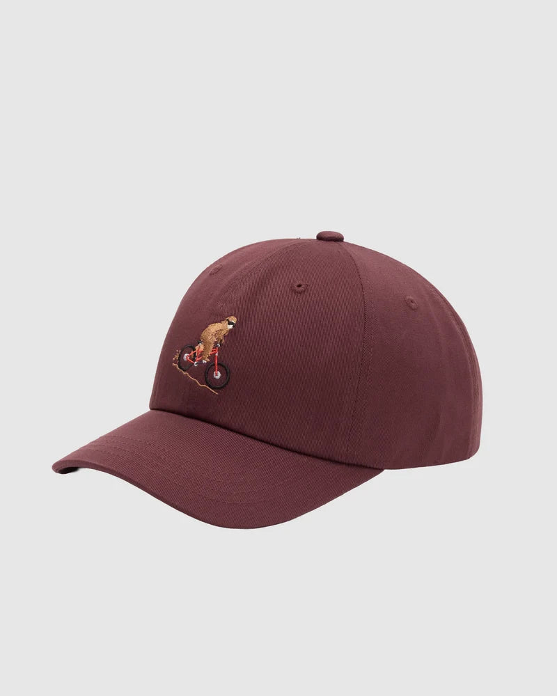 Tentree Sasquatch Peak Hat A maroon baseball cap featuring an embroidered graphic of a monkey riding a bicycle on the front, set against a solid light gray background.