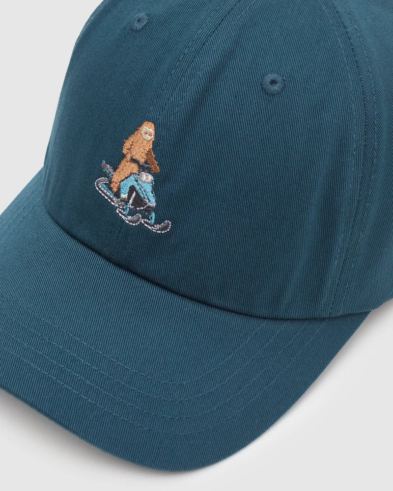 Tentree Sasquatch Peak Hat A close-up of a teal blue baseball cap featuring an embroidered graphic of a brown furry figure riding a light blue snowmobile.