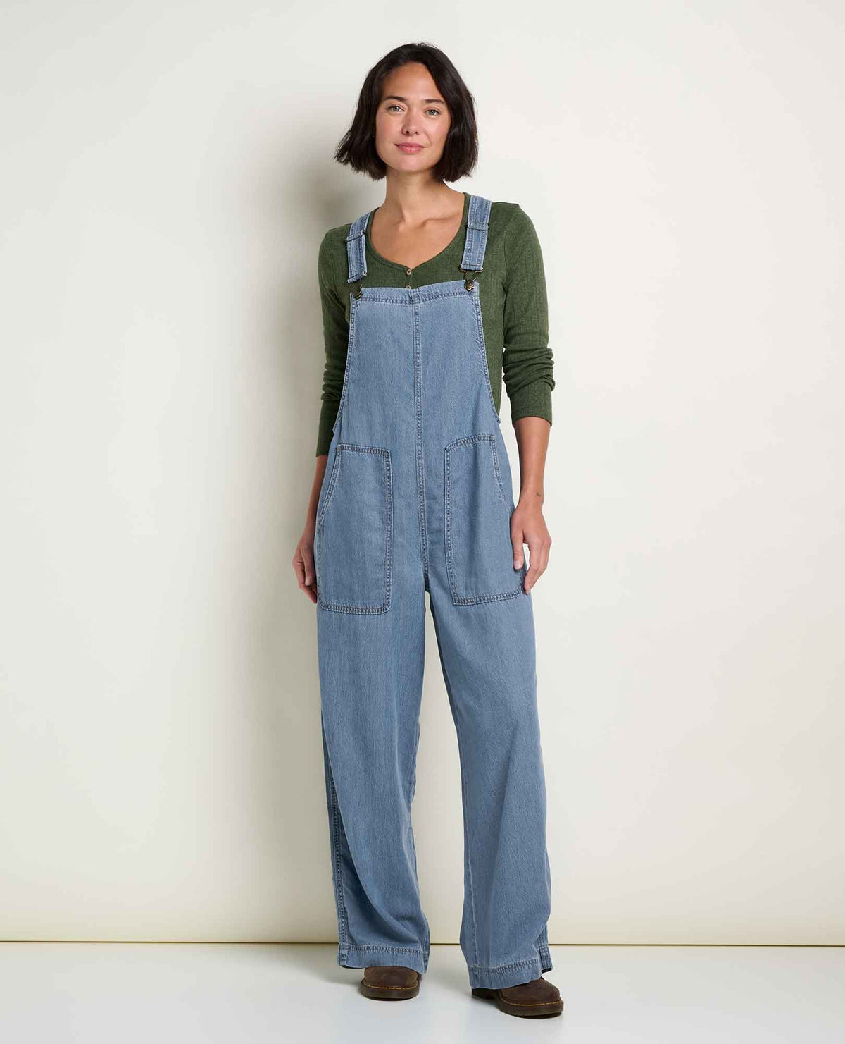 A woman stands with blue overalls on and a green long sleeve underneath.