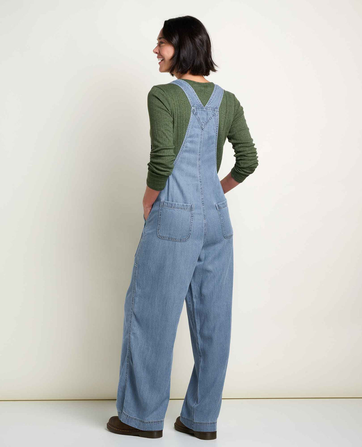 The reverse view of a woman standing with blue overalls and a green long sleeve shirt.