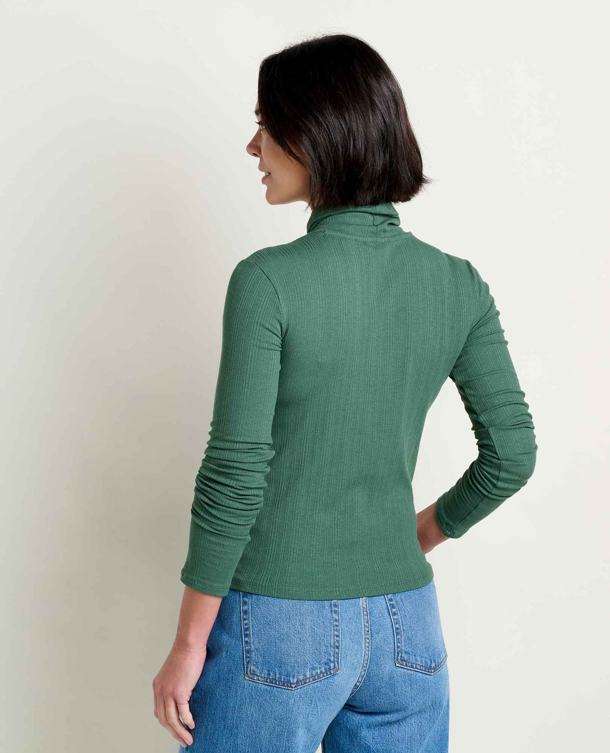 Toad & Co Rosalie Long Sleeve Turtle Neck A back view of a woman wearing a ribbed green turtlenecks and jeans with back pockets.