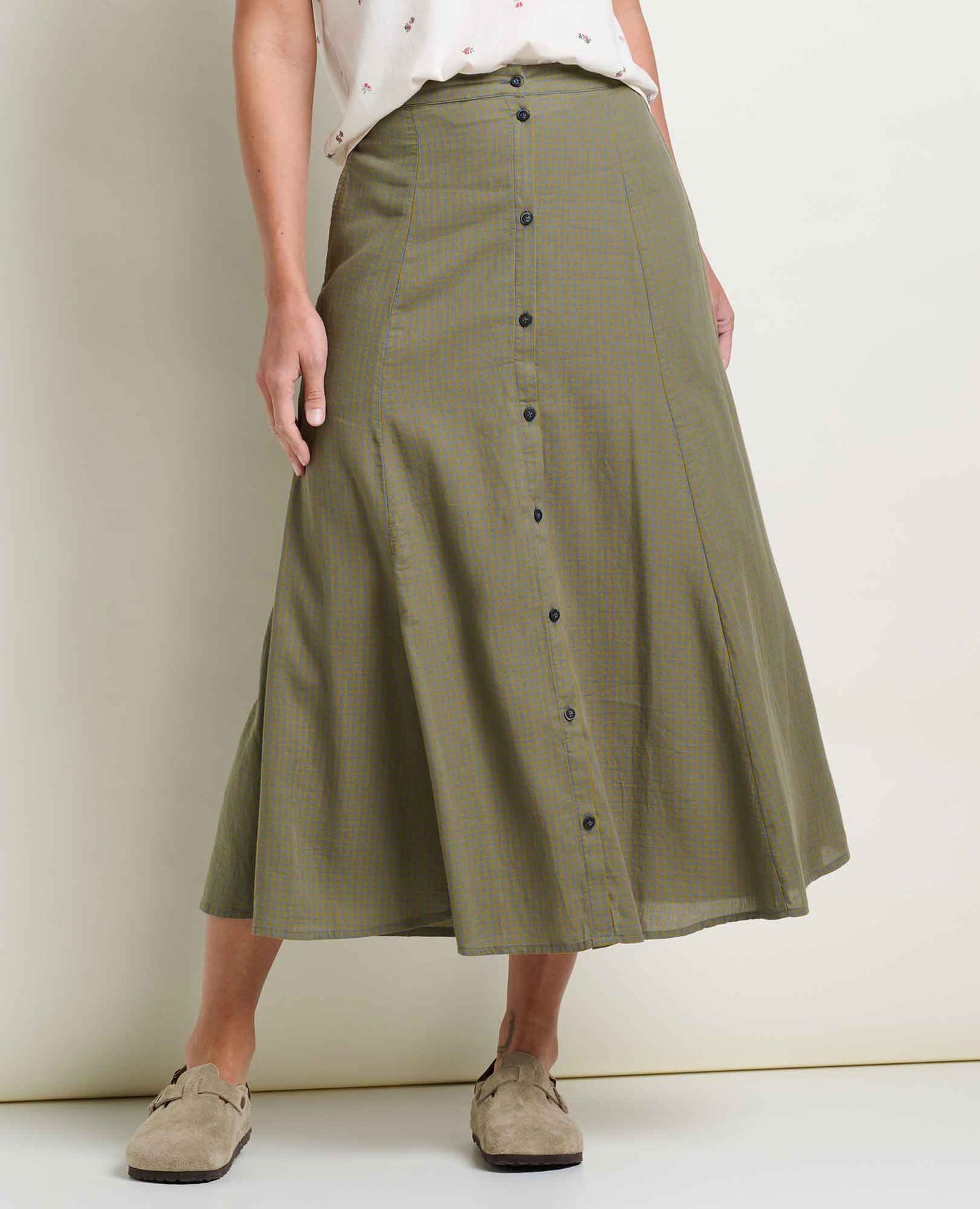 Toad and Co Manzana Organic Cotton Midi Skirt Green Moss Pattern A model wearing an olive green and beige checkered midi skirt with a button-down front, styled with a white top and beige suede loafers against a plain cream wall.