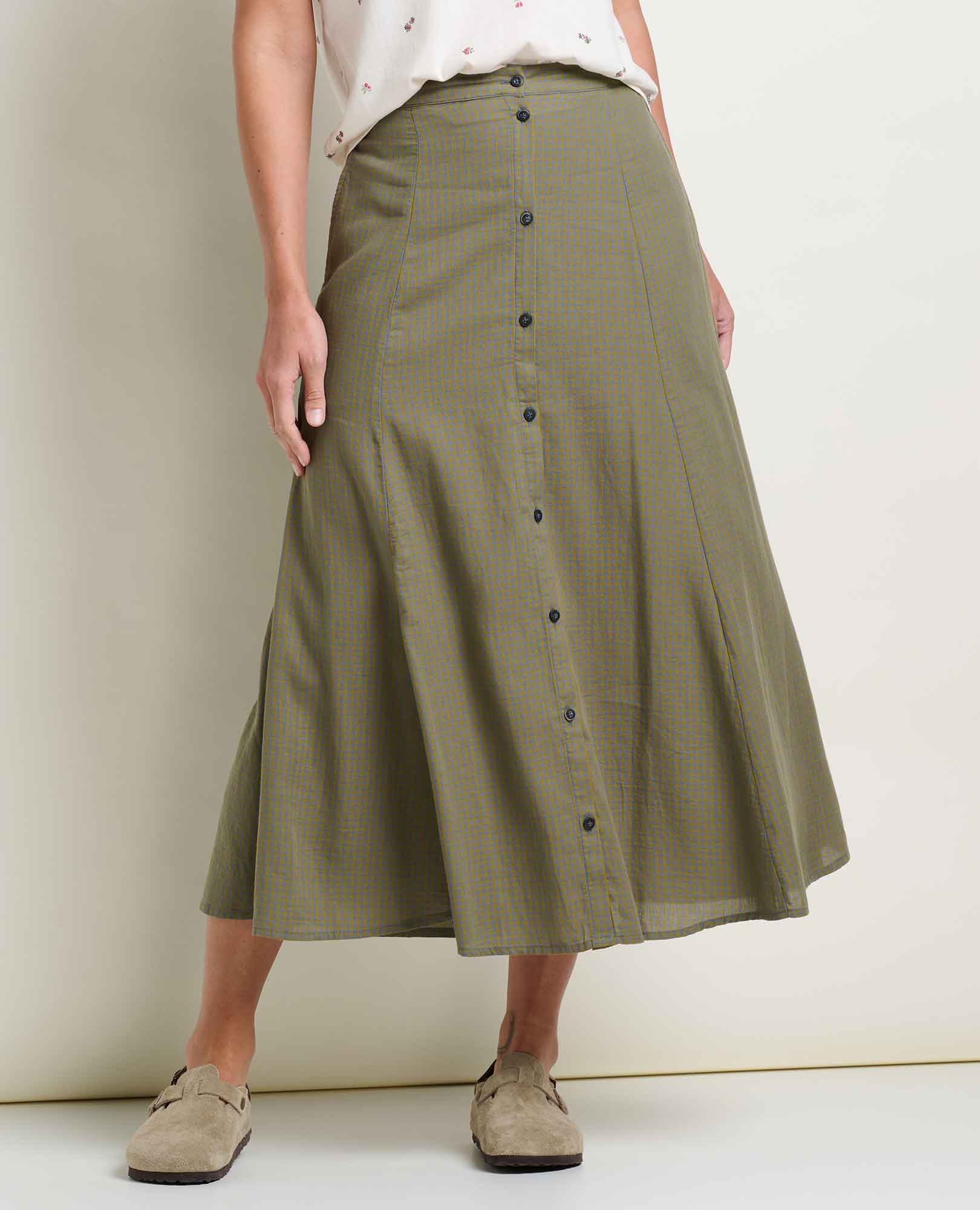 Toad and Co Manzana Organic Cotton Midi Skirt Green Moss Pattern A model wearing an olive green and beige checkered midi skirt with a button-down front, styled with a white top and beige suede loafers against a plain cream wall.