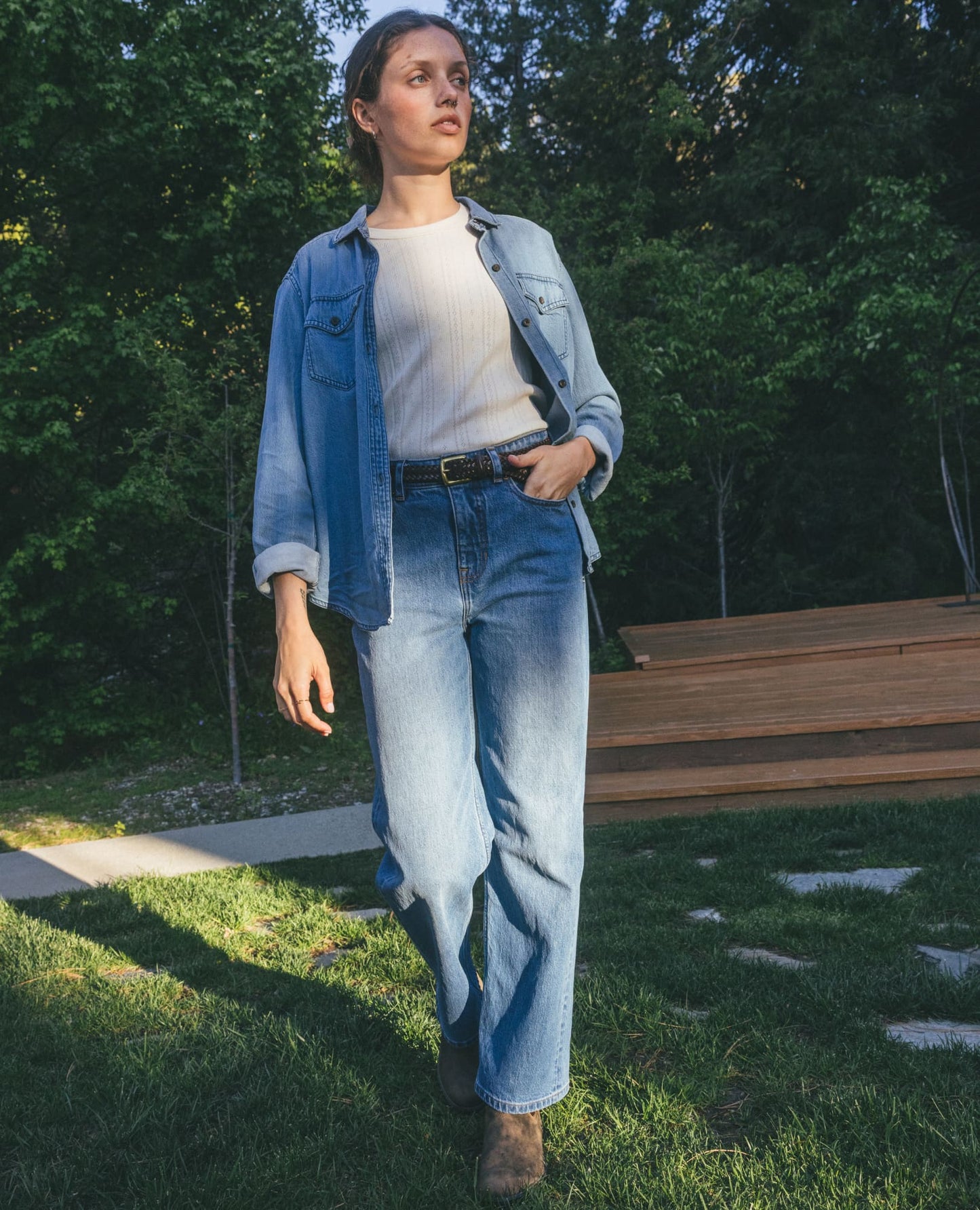 Toad and Co Tamarack Wide leg Jean A woman stands outdoors on grass wearing a light blue denim jacket over a cream ribbed top and light blue straight-leg jeans, with brown boots, against a background of green trees and a wooden bench.