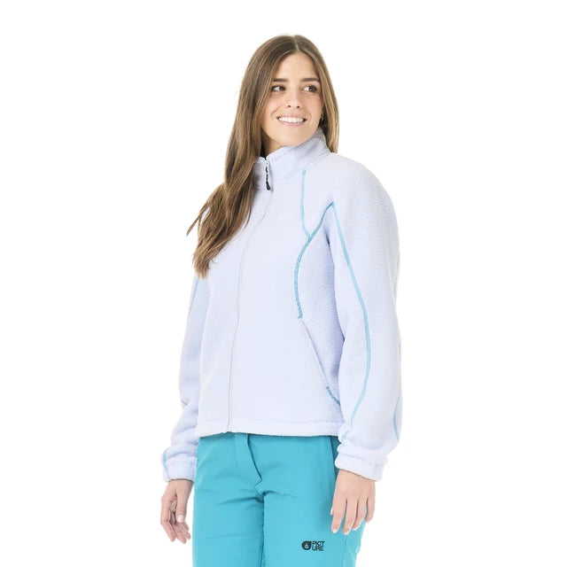 A woman stands in a white studio space wearing a light blue full zip sweater and blue pants