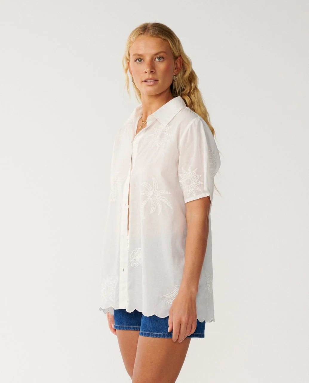 Rip Curl Bossa Nova Emb Short Sleeve Shirt A blonde model wearing a white short-sleeve button-up shirt with delicate floral embroidery and a scalloped hem, paired with blue denim shorts, against a plain light gray studio background.