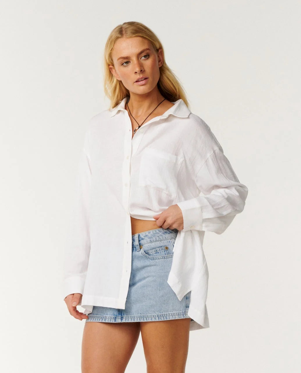 rip curl vibe A blonde model wearing a white long-sleeve button-up shirt and a light blue denim mini skirt, standing against a plain white studio background.